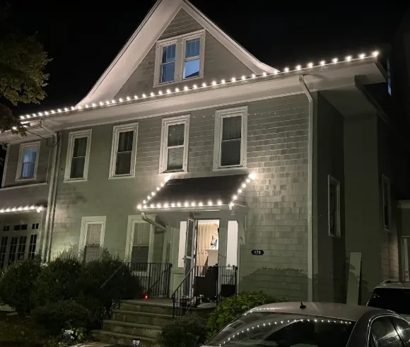 Shingle-style home with warm white lights along every eave and the porch roof
